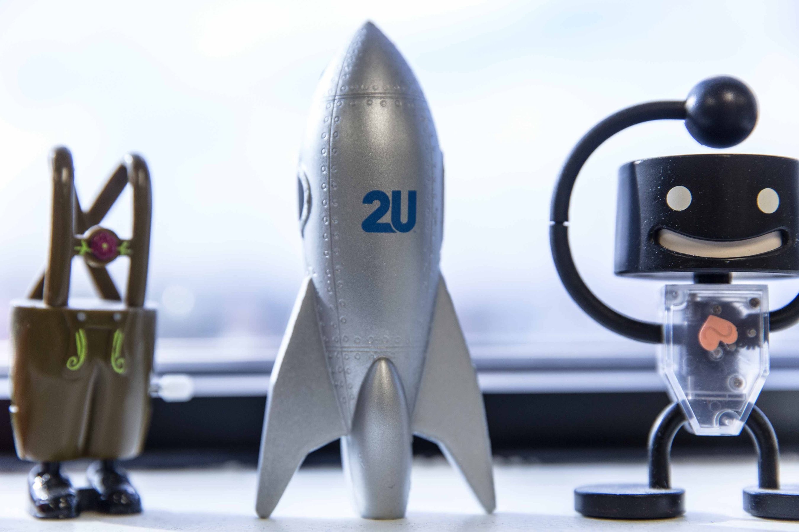 A silver rocket with the 2U logo on it sits in between a smiling robot toy and an overalls with feet toy