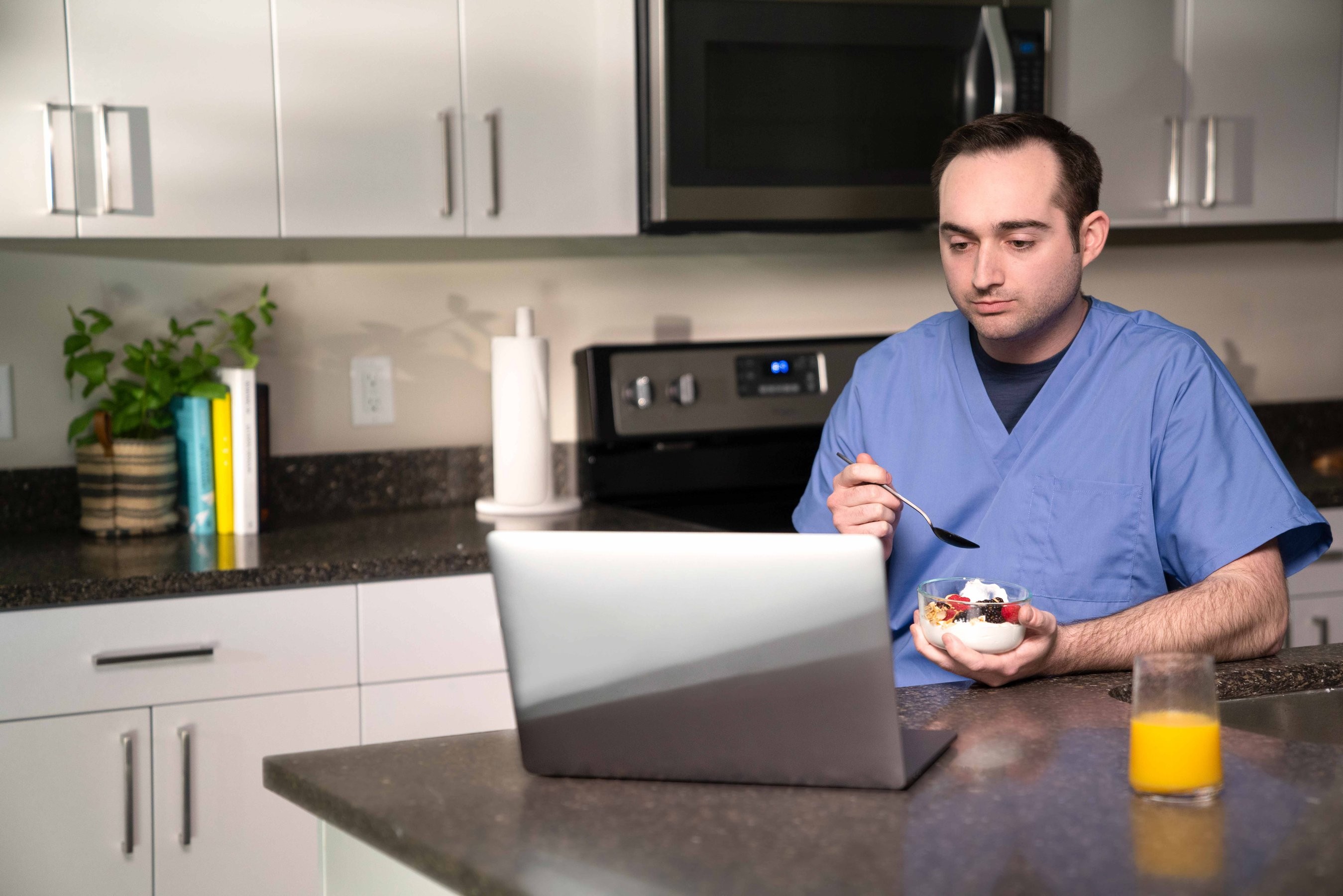 A man wearing a nurse scrub eats yogurt in his kitchen while looking at his laptop
