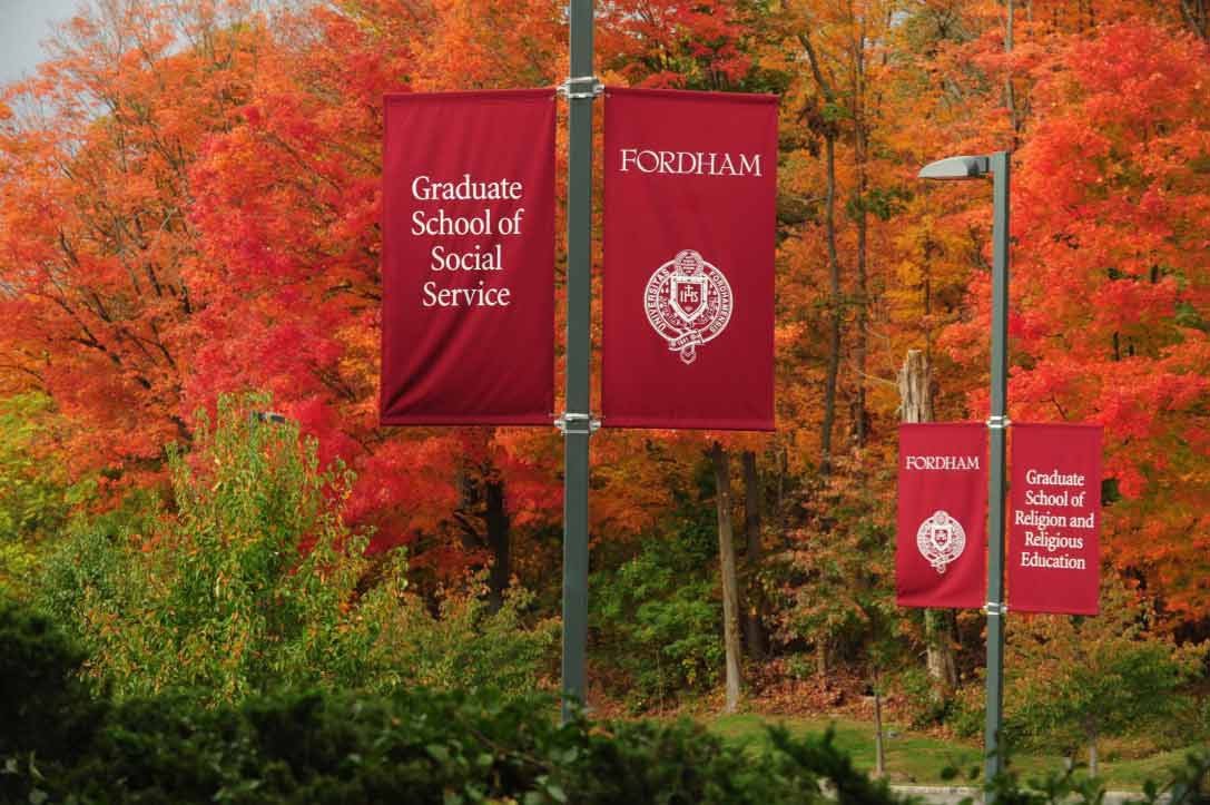 Red banners for Fordham University Graduate School of Social Service line a wooded walkway next to trees during fall