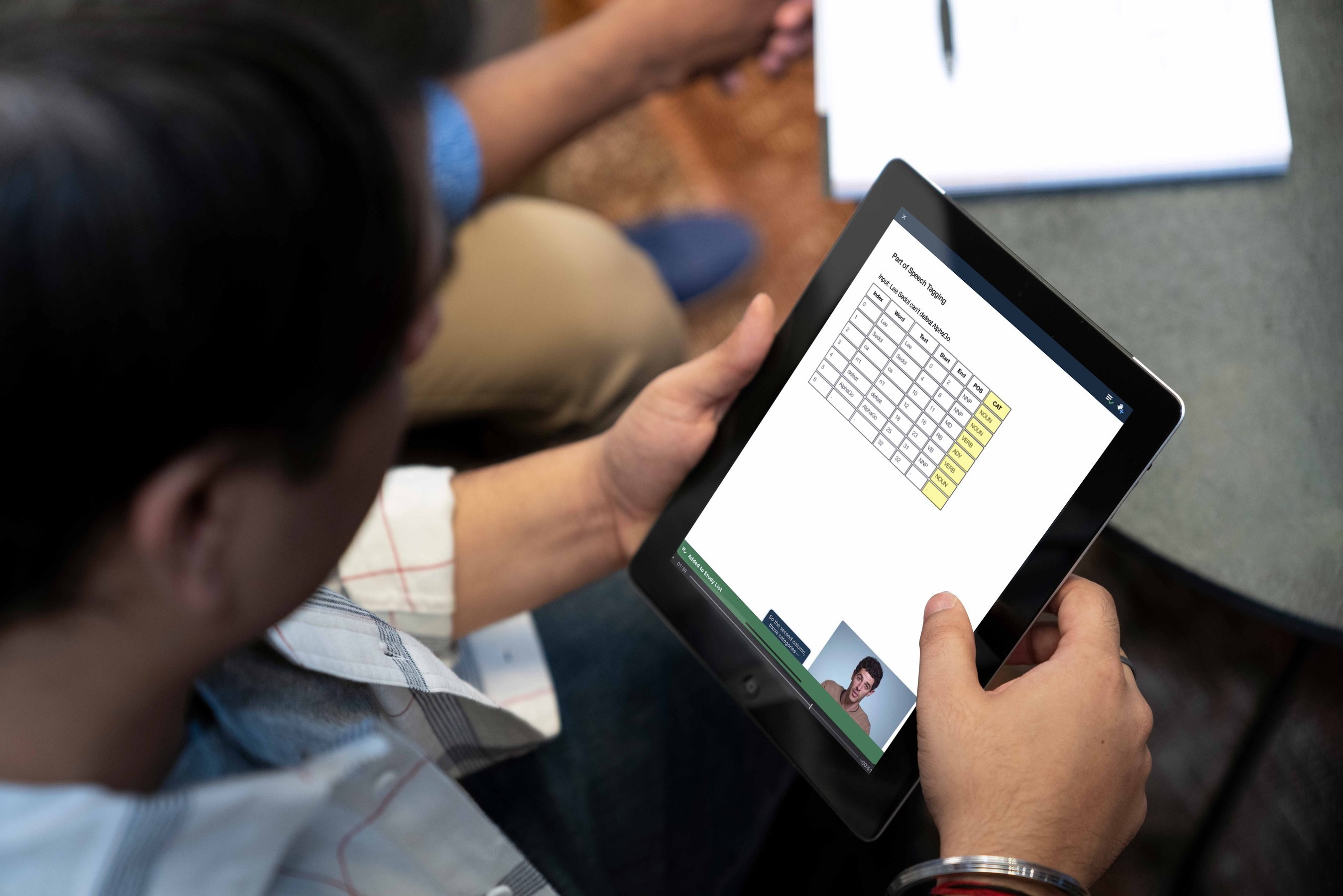 A man looking down at a tablet device with a data table on the screen and another man talking in a video thumbnail