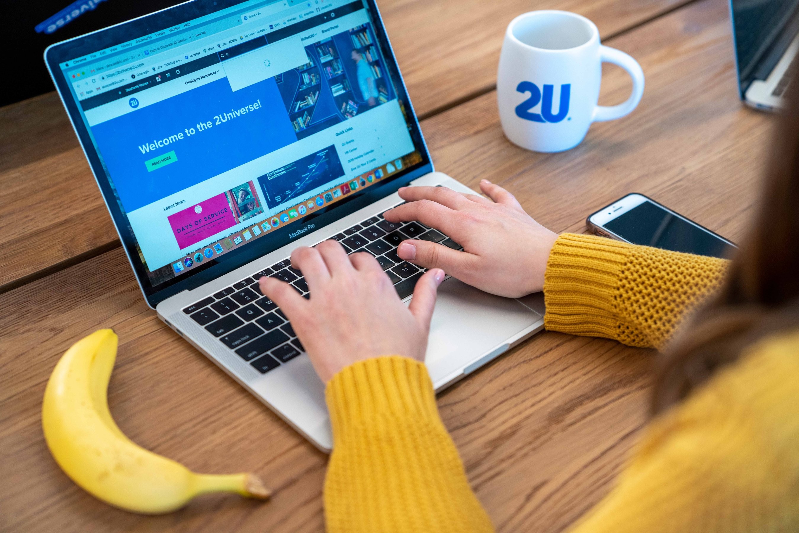 Woman on her laptop browses the 2Universe website with a 2U coffee mug and banana next to her